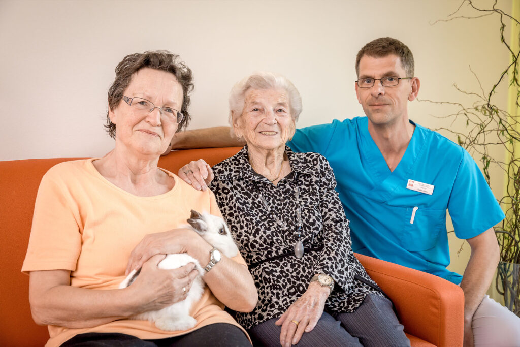 Betreuer kümmert sich um zwei Seniorinnen auf einem Sofa